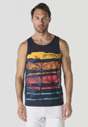 Hombre vistiendo una camiseta sin mangas azul marino con rayas horizontales de colores de atardecer y estampados de palmeras, combinado con pantalones blancos, de pie frente a un fondo liso.