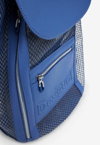 Blauer Rucksack mit einer glatten Lederklappe, Netzseiten, einer Front-Reißverschlusstasche und markenspezifischen strukturierten Details. Die Reißverschlüsse haben silberne Hardware-Akzente.