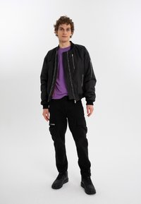 Black bomber jacket with zipper pockets, paired with a purple t-shirt and black cargo pants featuring side pockets and logo. Black sneakers.