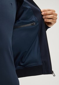 Marineblauwe zip-jack heeft aan de binnenkant een gladde stof met een ritszak. Ribbelboorden en een ribbelzoom verbeteren de pasvorm en structuur.