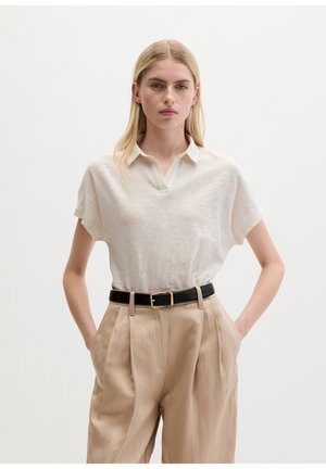 Femme aux cheveux blonds portant une chemise blanche à manches courtes avec col, un pantalon plissé beige taille haute et une ceinture noire avec une boucle dorée, les mains dans les poches.