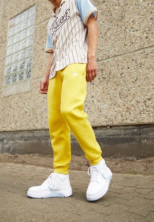 Personne portant un pantalon de survêtement Nike jaune, des baskets blanches montantes et un t-shirt beige style baseball avec des manches bleues, debout sur un trottoir pavé.