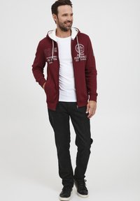 Felpa burgundy con zip e dettagli bianchi, caratterizzata da grafiche testuali, indossata sopra una t-shirt bianca con jeans neri e sneakers nere.