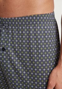 Shorts gris foncé avec un motif géométrique de croix blanches et vertes. La taille est dotée d'un bouton et du nom de la marque "CALIDA."