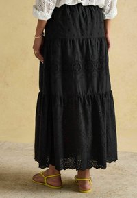 Czarna długa spódnica maxi z haftem oczkowym, warstwowymi falbanami i ząbkowanym dołem. Wygodne dopasowanie i lekka tkanina. Noszona z żółtymi sandałami.