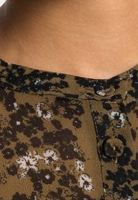 Brun blommönstrad blus med rund halsringning och två knappar. Mjuk tyg med en texturerad yta och svarta detaljer.