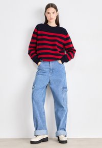 Pulover cu dungi roșii și bleumarin, cu guler rotund, asortat cu blugi cargo largi, de culoare bleu deschis, și pantofi bej cu șireturi, având talpa neagră.