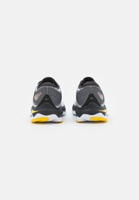 Zapatillas deportivas negras y grises con parte superior de malla texturada, que presentan una suela intermedia blanca y acentos en amarillo y azul en la suela exterior.
