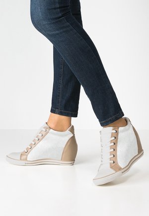 Person wearing white and beige wedge sneakers with laces and dark blue skinny jeans, standing on a white surface.