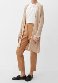 Cardigan beige a maglia su una maglietta bianca, abbinato a pantaloni color tan e mocassini neri. Il cardigan presenta polsini a coste e una apertura frontale.