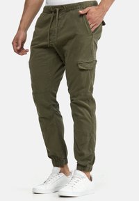 Pantaloni cargo verde măsliniu cu talie elastică, șnur, buzunar lateral cargo, picioare conice și tivuri elastice. Purtați cu teniși albi.