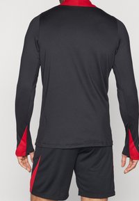 Langärmliges Sportshirt in Schwarz mit roten Akzenten, ausgestattet mit einem hohen Kragen und Daumenlöchern. Das Material wirkt glatt und dehnbar.