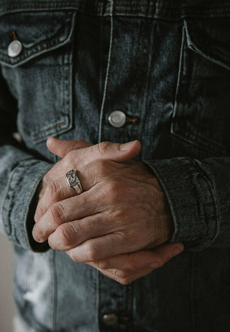 Jeansjacka med metallknappar, händerna hopknäppta, bär en silverring med distinkt detaljer. Strukturerad tyg och sliten utseende.