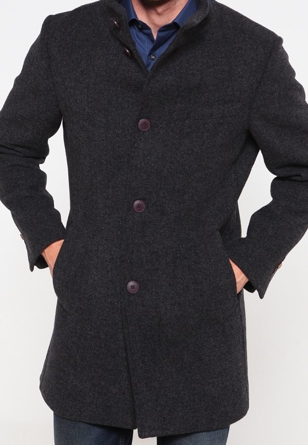 Homme portant un manteau en laine gris foncé à trois boutons, les mains dans les poches, par-dessus une chemise bleue à col et un jean.