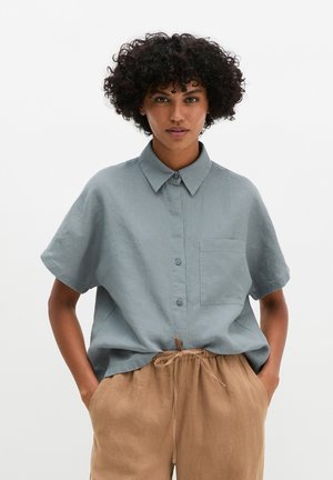Woman with curly hair wearing a loose blue short-sleeve button-up shirt and beige drawstring pants, standing with hands in pockets.