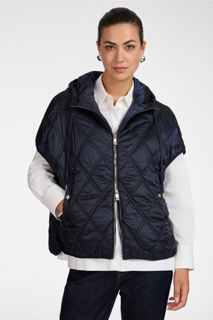 Frau trägt eine marineblaue gesteppte ärmellose Jacke über einem weißen Hemd mit Knöpfen und dunklen Jeans, steht vor einem schlichten Hintergrund.