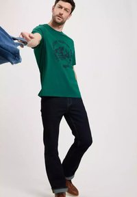 Zielona bawełniana koszulka z nadrukiem graficznym, połączona z ciemnoniebieskimi dżinsami i brązowymi butami, na jednolitym jasnym tle.