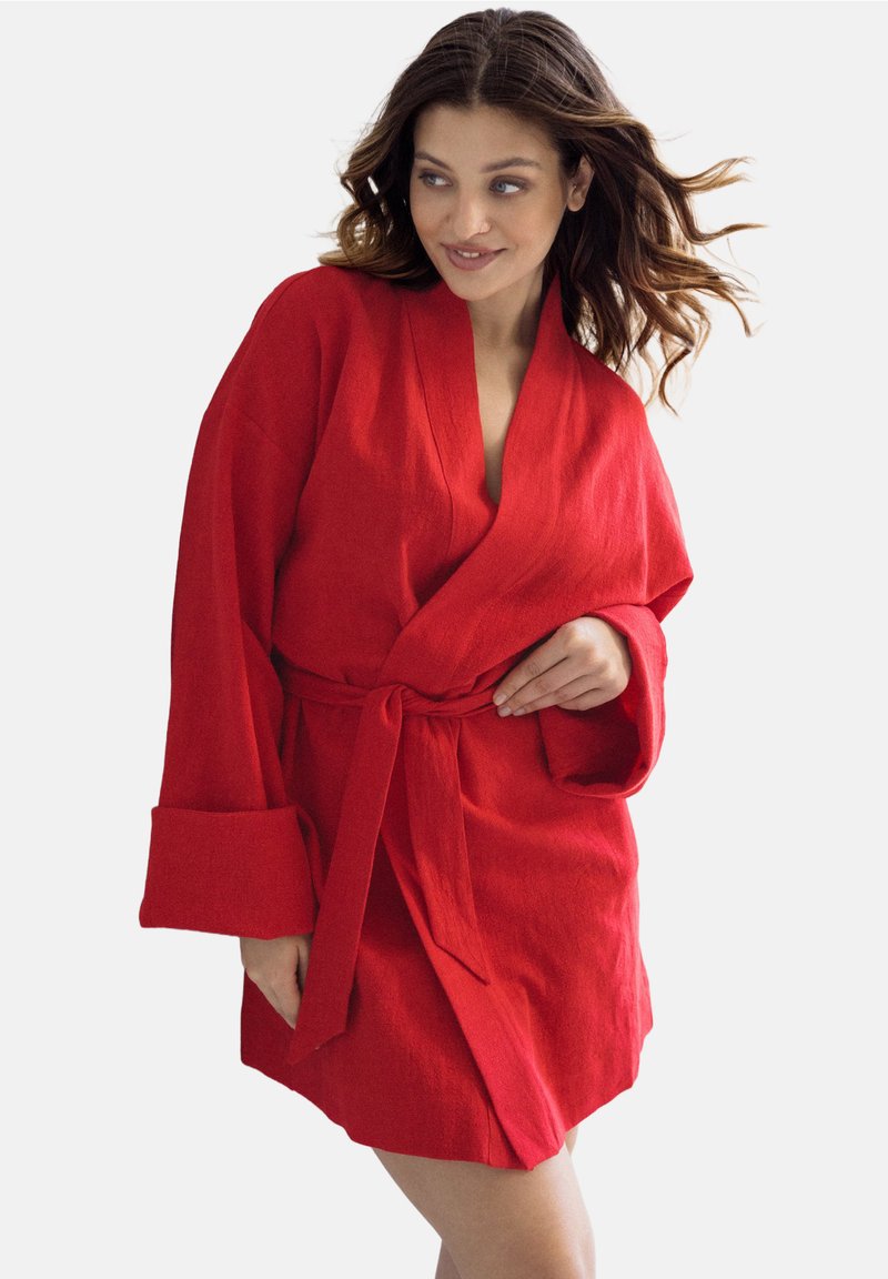 Czerwona lniana szata z szerokimi rękawami i pasek w talii. Charakteryzuje się dekoltem w kształcie litery V oraz teksturowaną tkaniną, zaprezentowana na modelce.