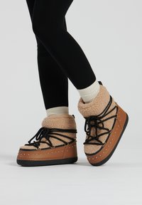 Botas de plataforma beige con parte superior de felpa, cordones negros y detalles de tachuelas a lo largo de la suela, combinadas con leggings negros y calcetines crema.