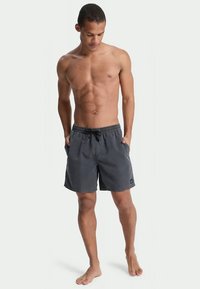 Pantaloni da bagno grigi con una vita elastica e lacci di regolazione, dotati di tasche laterali e una patch con logo sulla gamba. Tessuto morbido.