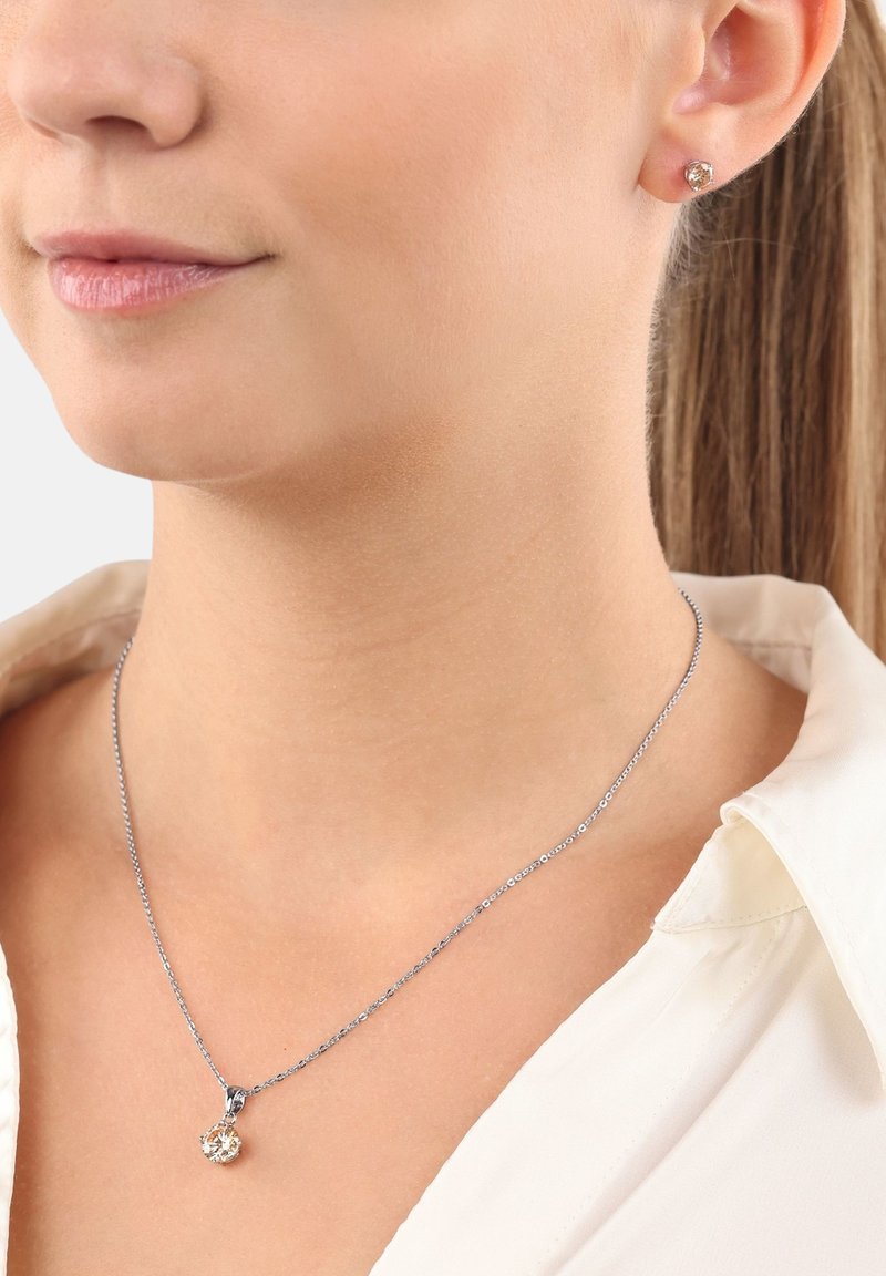 Collar de plata con un colgante redondo y pendientes de botón a juego. Ambos cuentan con piedras preciosas transparentes, destacando un diseño simple y elegante.