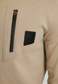 Pullover beige con tasca sul petto con zip. Caratterizzato da una texture liscia, un accento in tessuto nero e un elemento di branding sottile.