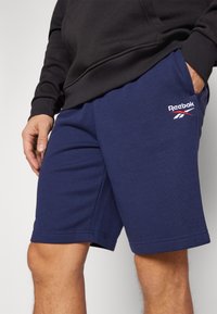 Plave kratke hlače od pamučne mješavine s elastičnom jeansom, bočnim džepovima i bijelim "Reebok" logom na lijevoj nozi. Glatka tekstura, do koljena.