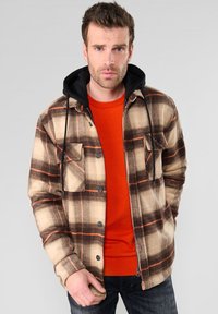 Veste à carreaux beige et marron avec des accents orange, dotée d'une capuche noire et de boutons. Portée par-dessus un pull orange vif.