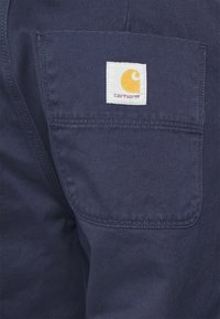 Marineblauwe werkbroek met een opgestikte zak en het Carhartt-logo, gemaakt van duurzaam materiaal met een gladde textuur en versterkte stiknaden.