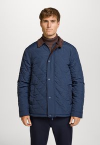 Chaqueta acolchada de color naval con cuello de ante marrón, que presenta cierres de botón y un corte relajado. Textura suave y patrón de diamantes en toda la prenda.