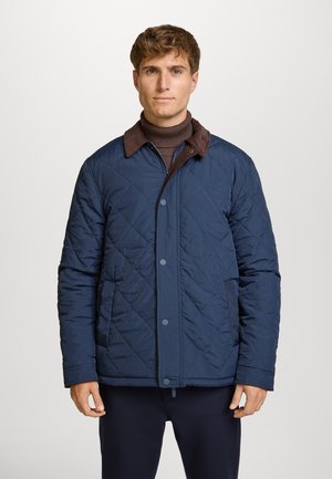 Chaqueta acolchada de color naval con cuello de ante marrón, que presenta cierres de botón y un corte relajado. Textura suave y patrón de diamantes en toda la prenda.