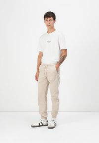 Beige jogger pants with elastic cuffs and drawstring waist paired with a white cotton t-shirt featuring a small logo. Black and white sneakers.