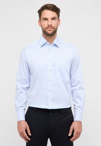 Chemise à manches longues rayée bleu clair et blanc avec col, boutons dorés et poche poitrine ; portée avec un pantalon noir.