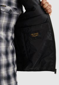 Hand hält eine offene schwarze Jacke, die das Etikett in der Innentasche mit der Aufschrift "PME Legend American Classic" zeigt, getragen über einem schwarz-weiß karierten Hemd.