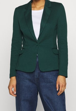 Persoon draagt een donkergroene blazer met één knoop over een wit overhemd en blauwe spijkerbroek, staand met ontspannen armen langs het lichaam.