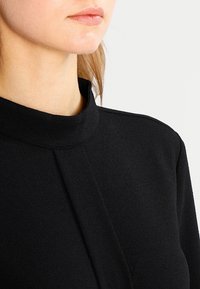 Indumento nero a collo alto realizzato in tessuto liscio, con una sofisticata cucitura lungo il davanti e spalle strutturate.