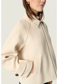 Beige ribbet jakke med en mock krave, lynlås lukning og sidelommer. Har en afslappet pasform og knapdetaljer ved ærmerne.