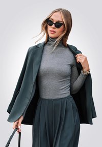 Donkergroene blazer over een grijze coltrui, gecombineerd met een geplooide grijze rok. Accessoires zijn onder andere een zonnebril en een handtasriem.