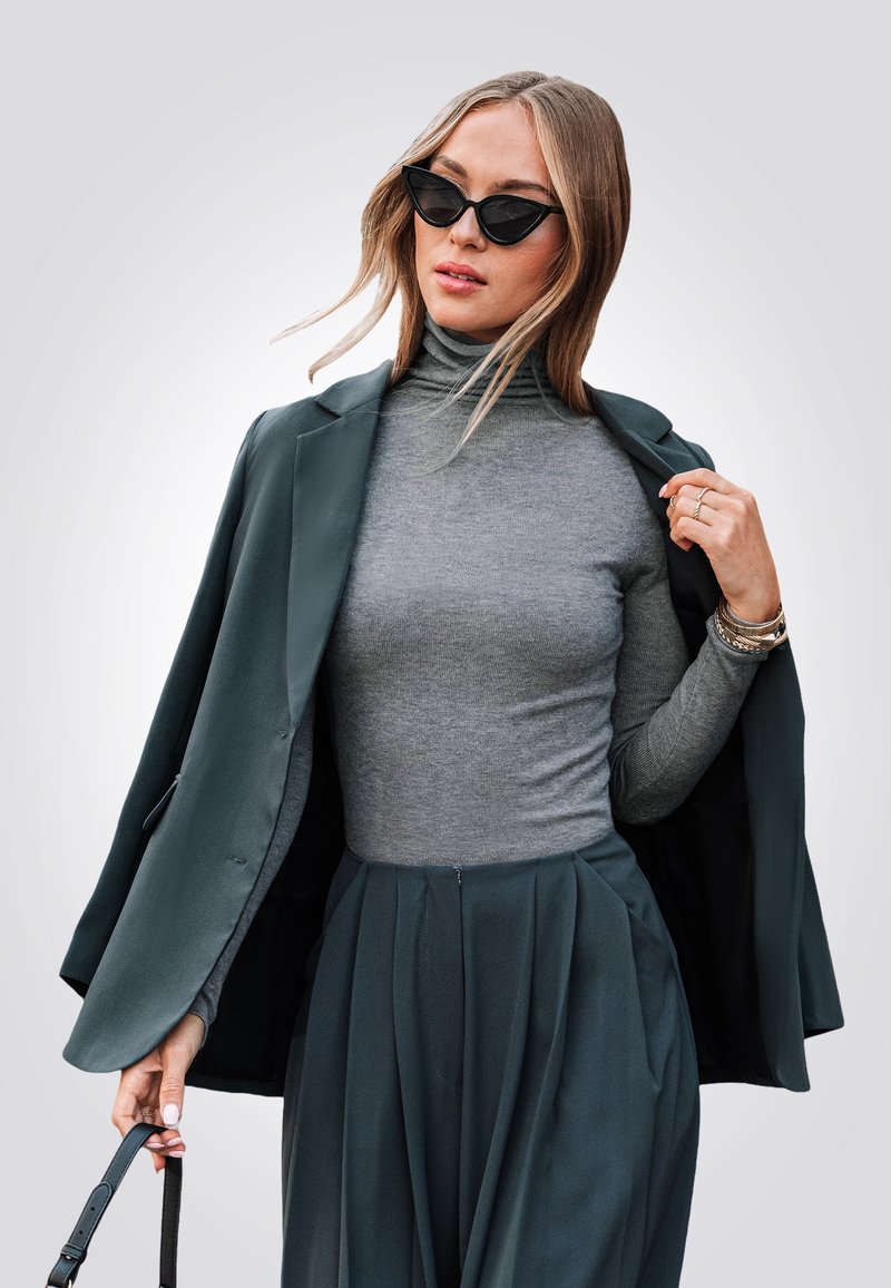 Donkergroene blazer over een grijze coltrui, gecombineerd met een geplooide grijze rok. Accessoires zijn onder andere een zonnebril en een handtasriem.