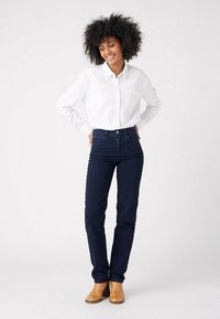 Donkerdenim jeans met een aansluitend ontwerp, hoge taille en subtiele stikdetails, gecombineerd met een wit blouse met knopen en tan enkellaarsjes.