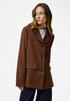 Noor naine laineliste blondide juustega, kellel on seljas pruun nööpidega mantel valge topi ja tumedate pükstega, vaatamas oma vasakule tavalise tausta ees.