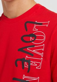Červené bavlněné tričko s výrazným černým textem "LOVE" vertikálně podél levé strany. Hladký povrch a klasický kulatý výstřih.