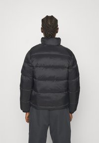 Personne aux cheveux tressés portant une doudoune noire et un pantalon sombre, vue de dos sur un fond clair uni.