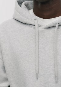 Harmaa huppari, valmistettu pehmeästä kankaasta, jossa on kiristysnauhalla varustettu huppu ja yksinkertainen muotoilu. Näkyvä sisäpuolen valkoinen paidan kaulus.