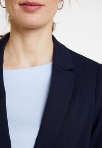 Granatowa marynarka z klapami, w dopasowanym fasonie, uszyta z teksturowanej tkaniny na jasnoniebieskiej bluzce. Widoczne złote kolczyki w kształcie kół.