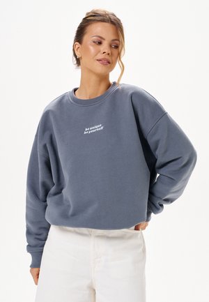 Femme à la peau claire et aux cheveux attachés, portant un sweat bleu avec le texte « be unique be yourself » et un pantalon blanc, debout avec une main sur la hanche.