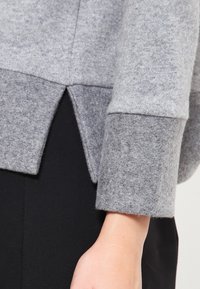 Szary sweter z teksturowanej tkaniny, z rozciętym dołem i kontrastującymi szarymi mankietami. Projekt obejmuje luźny krój i miękki materiał.