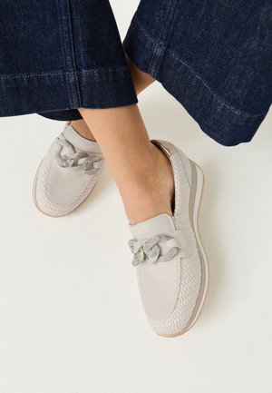 Kvinde iført hvide slip-on vævede loafers med chunky kædedetalje, kombineret med mørkeblå cropped denim bukser.