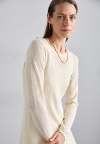Maglione lavorato a maglia color crema con un design a rete, con maniche lunghe e scollatura rotonda. Collana in oro e orecchini a cerchio visibili.