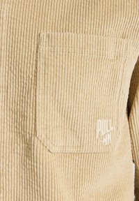 Chemise en velours côtelé beige avec une poche appliquée et un logo embossé. Le tissu présente un motif côtelé texturé.